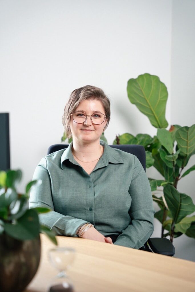Mitarbeiterin Nelli Giesbrecht sitzt lächelnd am Schreibtisch mit großer Zimmerpflanze im Hintergrund
